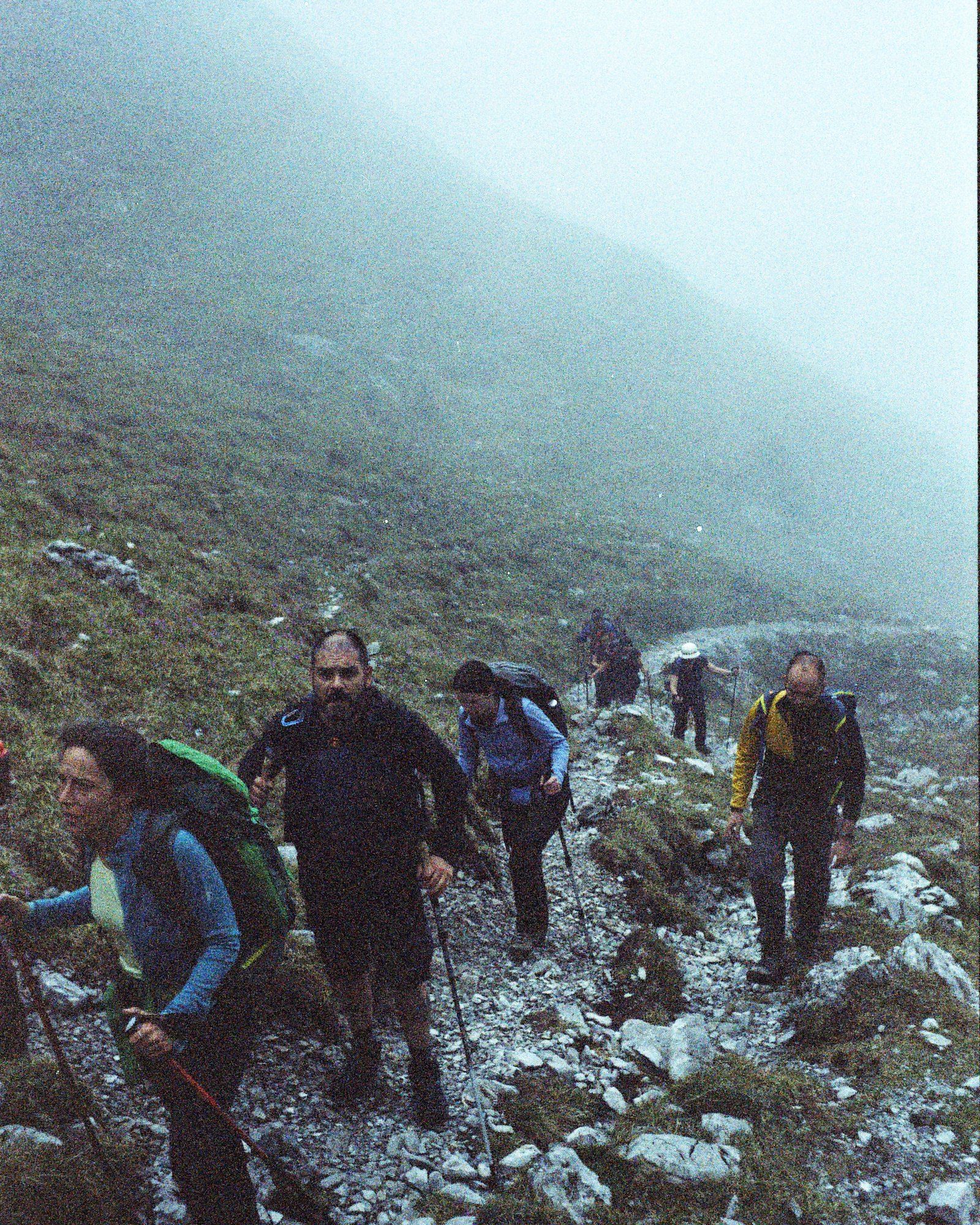 Il gruppo sale con i bastoncini su un pendio di sassi e prato, con la montagna nella nebbia
