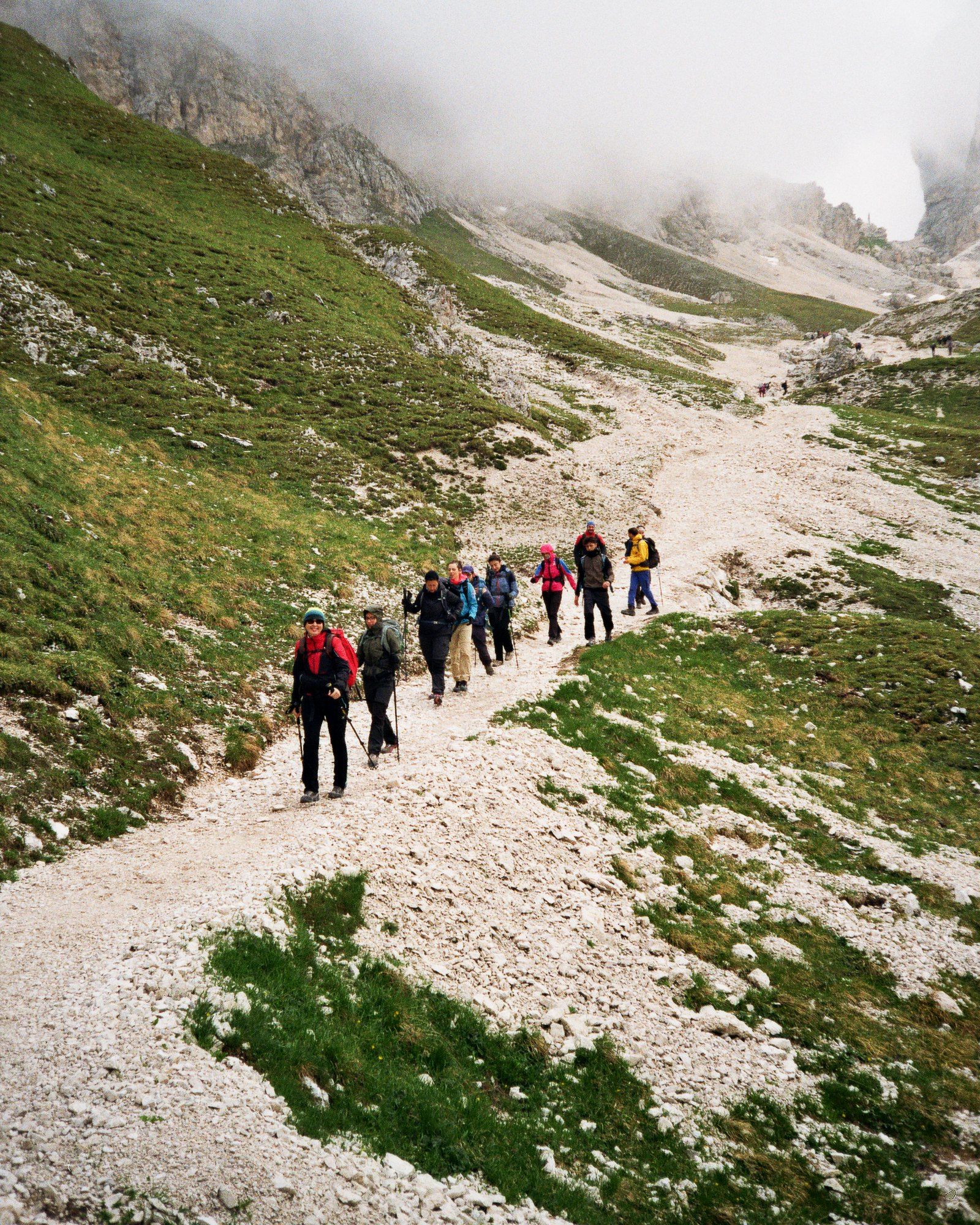 Una fila di escursionisti sale su un sentiero di ghiaia chiara in un vallone delle Dolomiti, con le pareti nella nebbia