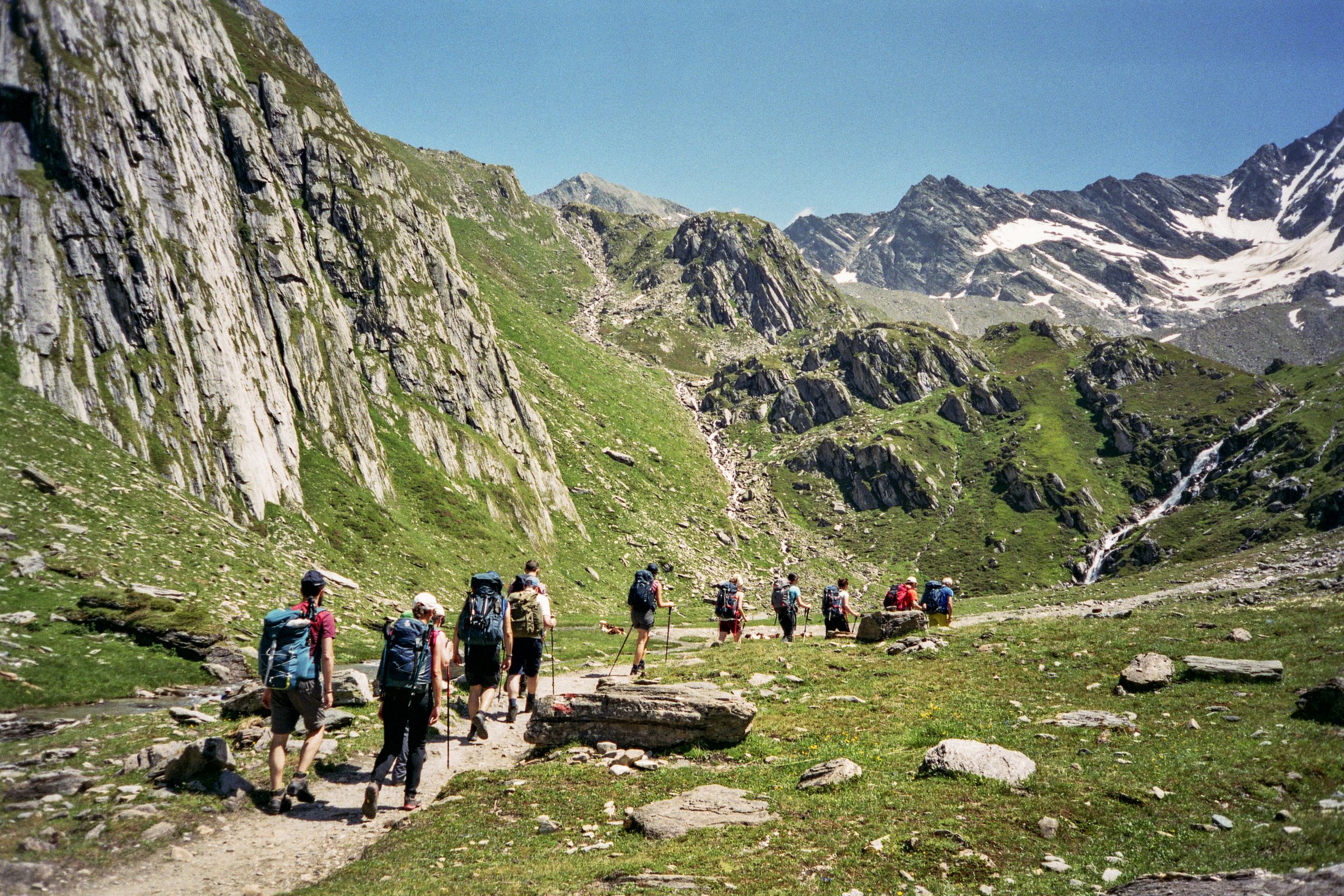Un gruppo di escursionisti in fila su un sentiero di fondovalle, tra prati e pareti di roccia